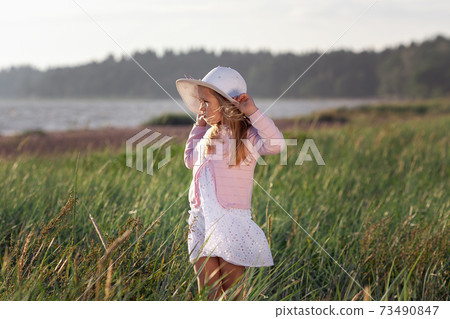 Cute little girl in white dress and hat looking to the sea among green field of grass in sunset Cute little girl in white dress and hat looking to the sea among green field of grass in sunset 73490847