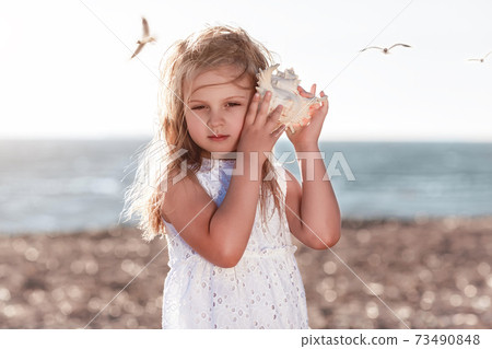 Little caucasian blond girl holding seashell and listening to the sea Little caucasian blond girl holding seashell and listening to the sea 73490848