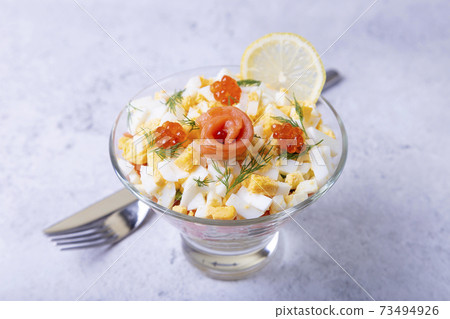 Layered salad with salted trout (salmon), caviar, eggs and vegetables. Traditional dish, one portion in a glass goblet on a gray background. Close-up. Layered salad with salted trout (salmon), caviar, eggs and vegetables. Traditional dish, one portion in a glass goblet on a gray background. Close-up. 73494926