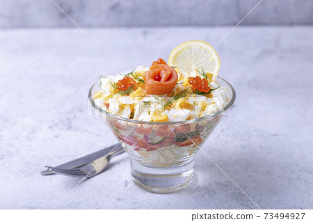 Layered salad with salted trout (salmon), caviar, eggs and vegetables. Traditional dish, one portion in a glass goblet on a gray background. Close-up. 73494927