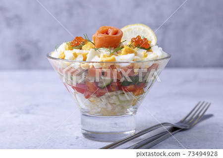 Layered salad with salted trout (salmon), caviar, eggs and vegetables. Traditional dish, one portion in a glass goblet on a gray background. Close-up. Layered salad with salted trout (salmon), caviar, eggs and vegetables. Traditional dish, one portion in a glass goblet on a gray background. Close-up. 73494928