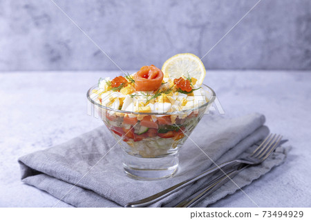 Layered salad with salted trout (salmon), caviar, eggs and vegetables. Traditional dish, one portion in a glass goblet on a gray background. Close-up. Layered salad with salted trout (salmon), caviar, eggs and vegetables. Traditional dish, one portion in a glass goblet on a gray background. Close-up. 73494929