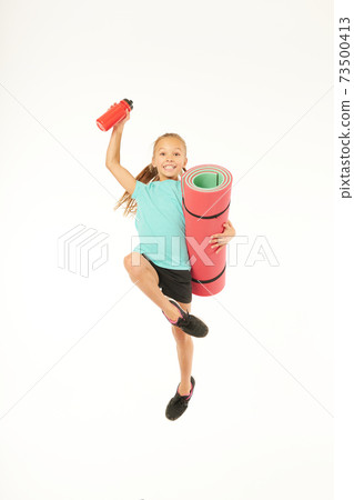 Joyful girl with yoga mat and bottle of water jumping in studio 73500413