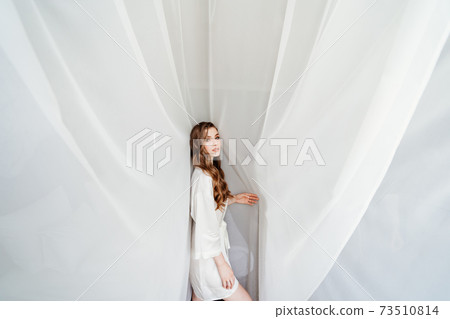 beautiful young woman with long hair in a white robe stands under a curtain beautiful young woman with long hair in a white robe stands under a curtain 73510814