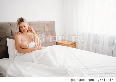 without clothes mother is sitting with a baby in a white bed feeding breast. without clothes mother is sitting with a baby in a white bed feeding breast. 73510817