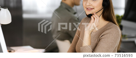 Young friendly girl in headsets is talking to a firm's client, while sitting at the desk in a modern office together with her colleague. Call center operators at work Young friendly girl in headsets is talking to a firm's client, while sitting at the desk in a modern office together with her colleague. Call center operators at work 73516593