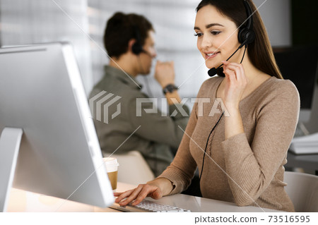 Young friendly girl in headsets is talking to a firm's client, while sitting at the desk in a modern office together with her colleague. Call center operators at work Young friendly girl in headsets is talking to a firm's client, while sitting at the desk in a modern office together with her colleague. Call center operators at work 73516595