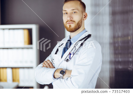 Red-bearded doctor standing straight in clinic near his working place. Portrait of physician. Medicine and healthcare concept Red-bearded doctor standing straight in clinic near his working place. Portrait of physician. Medicine and healthcare concept 73517403