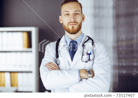 Red-bearded doctor standing straight in clinic near his working place. Portrait of physician. Medicine and healthcare concept Red-bearded doctor standing straight in clinic near his working place. Portrait of physician. Medicine and healthcare concept 73517407
