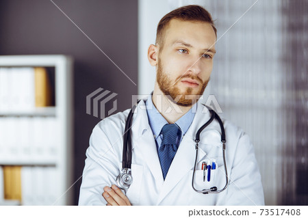 Red-bearded doctor standing straight in clinic near his working place. Portrait of physician. Medicine and healthcare concept Red-bearded doctor standing straight in clinic near his working place. Portrait of physician. Medicine and healthcare concept 73517408
