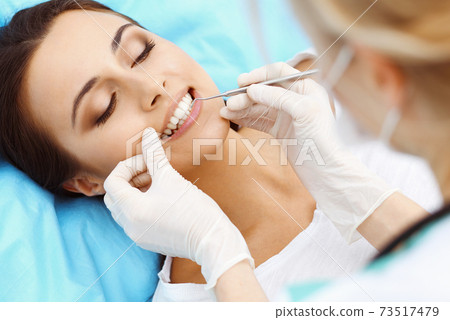 Young female patient visiting dentist office.Beautiful woman with healthy straight white teeth sitting at dental chair with open mouth during oral checkup while doctor working at teeth Young female patient visiting dentist office.Beautiful woman with healthy straight white teeth sitting at dental chair with open mouth during oral checkup while doctor working at teeth 73517479