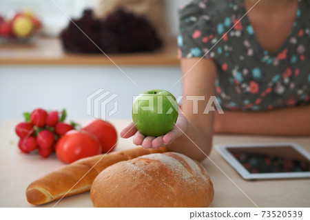 Close-up of female hand holding green apple in kitchen interiors. Many vegetables and other meal at glass table are ready for been cooked soon 73520539
