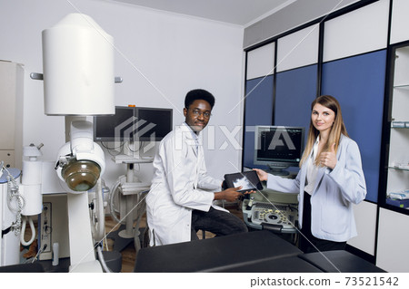 Portrait of smiling African man doctor, holding digital tablet with ultrasound scan, posing to camera, while consulting his female patient in modern clinic. Woman patient shows thumb up 73521542