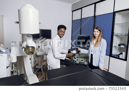 Portrait of smiling pretty woman, posing to camera, while visiting her African man doctor in modern clinic, holding digital tablet with ultrasound scan. Lithotripsy therapy and ultrasound Portrait of smiling pretty woman, posing to camera, while visiting her African man doctor in modern clinic, holding digital tablet with ultrasound scan. Lithotripsy therapy and ultrasound 73521543