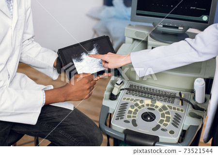 Cropped top view of black male doctor hands, holding ipad pc with ultrasound scan, while female hand of patient or doctor colleague pointing on tablet, on the background of modern ultrasound machine 73521544