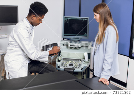 Young male african doctor in modern clinic, holding tablet pc and showing to his female caucasian patient ultrasound scan of kidneys and internal organs and explaining ways of treatment. Young male african doctor in modern clinic, holding tablet pc and showing to his female caucasian patient ultrasound scan of kidneys and internal organs and explaining ways of treatment. 73521562