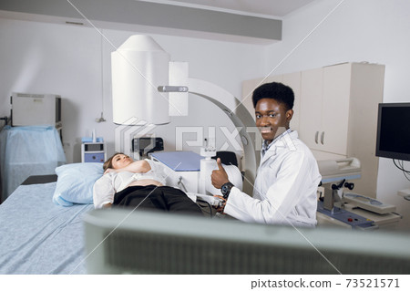 Portrait of young handsome African man doctor, looking at camera with thumb up, while providing lithotripsy procedure and ultrasound scan for his female Caucasian patient with modern lithotriptor Portrait of young handsome African man doctor, looking at camera with thumb up, while providing lithotripsy procedure and ultrasound scan for his female Caucasian patient with modern lithotriptor 73521571