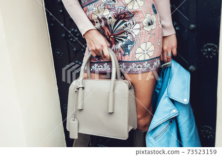 Autumn fashion image of young woman posing near black metal door, blue leather jacket, colourfull skirt and beige high autumn boots. Fashionable woman posing with beige handbag 73523159
