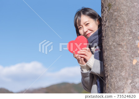 A woman holding a box of hearts under the blue sky and popping out from behind a tree 73524620