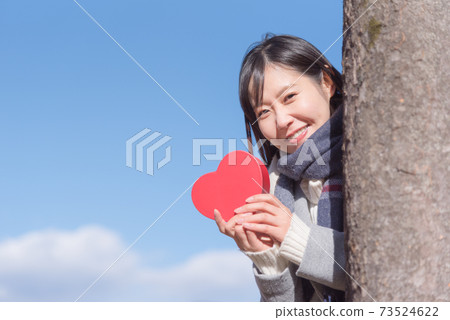 A woman holding a box of hearts under the blue sky and popping out from behind a tree 73524622