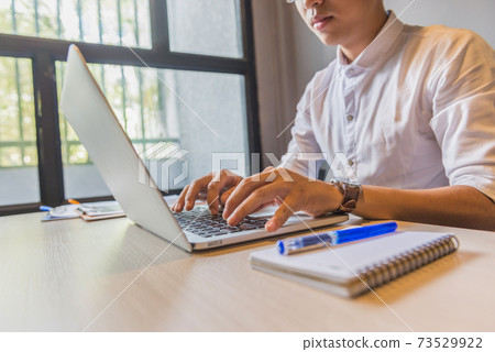 Asian man in white shirt typing laptop next to window Asian man in white shirt typing laptop next to window 73529922