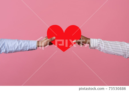 Cropped view of black man and his girlfriend holding red paper heart over pink studio background. Valentine's Day 73530886
