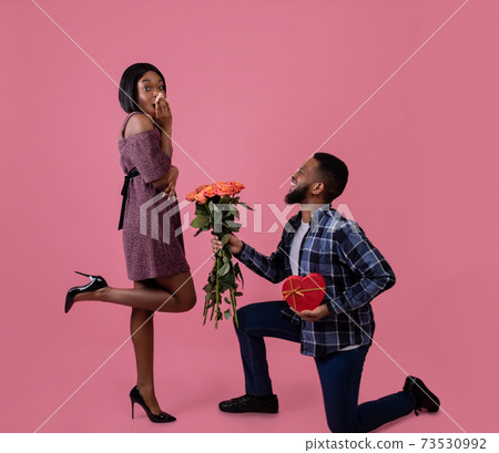 Passionate black man giving his sweetheart bouquet of roses and heart shaped gift for Valentine's Day on pink background 73530992