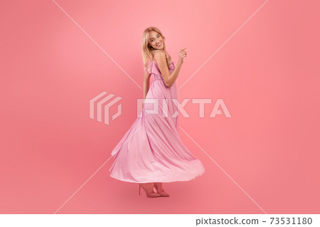 Fashionable blonde woman in long silk prom dress spinning around over pink background, copy space. Holiday concept Fashionable blonde woman in long silk prom dress spinning around over pink background, copy space. Holiday concept 73531180