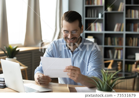 Smiling young businessman reading paper correspondence at home office. Smiling young businessman reading paper correspondence at home office. 73531679