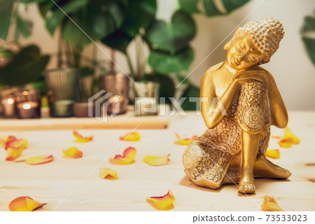 Golden bronze Buddha decorative statuette with flower petals on wooden table with burnig candles, monstera plant background. Meditation ritual, Spirituality, buddhism and religion. Selective focus Golden bronze Buddha decorative statuette with flower petals on wooden table with burnig candles, monstera plant background. Meditation ritual, Spirituality, buddhism and religion. Selective focus 73533023