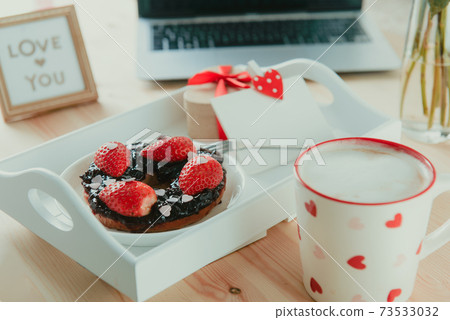 A tray with cup of latte coffee, bagel with strawberries and candy hearts, gift box and blank greeting card, Love you message on working place with laptop. Surprise for Valentine day. Selective focus. 73533032