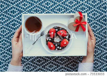 Top view female hands holding romantic meal for Lover. A cup of tea, bagel with jam decorated with strawberries and heart-shaped gift box on the white wooden tray. Simple surprise for Valentine's day Top view female hands holding romantic meal for Lover. A cup of tea, bagel with jam decorated with strawberries and heart-shaped gift box on the white wooden tray. Simple surprise for Valentine's day 73533033