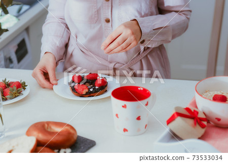 Woman decorating sweet bagel with jam with strawberries and heart-shaped candies. Preparing surprise breakfast for lover on Valentines' day. Simple festive food ideas. Copy space 73533034