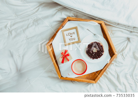 Top view homemade Valentine's day breakfast in bed for Lover. A cup of coffee and bagel with jam, heart-shaped gift box with red ribbon, Love you card on a wooden tray. Selective focus, copy space. 73533035