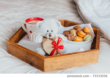 Valentine's day breakfast in bed for Lover. Teddy Bear with a heart-shaped gift box with red ribbon, a cup of coffee or cocoa and macaroons cookies on the wooden tray. Romantic morning. Copy space 73533036