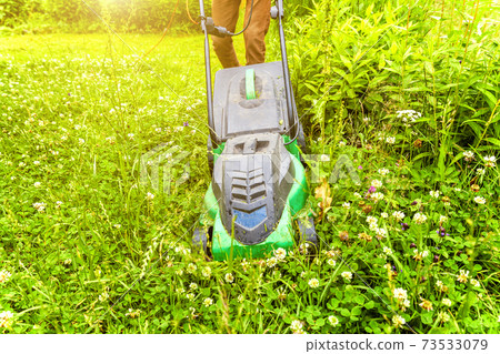 Man cutting green grass with lawn mower in backyard. Gardening country lifestyle background. Beautiful view on fresh green grass lawn in sunlight, garden landscape in spring or summer season. Man cutting green grass with lawn mower in backyard. Gardening country lifestyle background. Beautiful view on fresh green grass lawn in sunlight, garden landscape in spring or summer season. 73533079