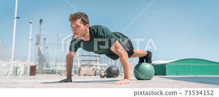 Fitness man banner panorama. Athlete strength training pushup balancing legs on medicine ball for advanced core body workout push-ups floor exercises at outdoor gym Fitness man banner panorama. Athlete strength training pushup balancing legs on medicine ball for advanced core body workout push-ups floor exercises at outdoor gym 73534152