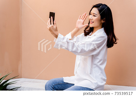 Young asian girl with bright red lipstick is taking a selfie with her smartphone having a desert sand colored wall in her background wearing a white shirt with open buttons and an open hairstyle 73539517