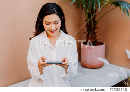 Asian woman playing games on smartphone while sitting on a concrete bench surrounded with pots of green plants with a desert sand colored wall in her background 73539526