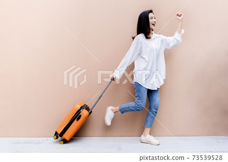 Young Asian girl happy while towing her bright orange suitcase with right hand and left hand fist bumping in the air on concrete floor and desert sand colored wall in her background 73539528