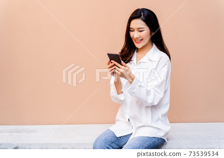 Asian woman texting on smartphone while sitting on a concrete bench surrounded with pots of green plants with a desert sand colored wall in her background and green leaves in the foreground Asian woman texting on smartphone while sitting on a concrete bench surrounded with pots of green plants with a desert sand colored wall in her background and green leaves in the foreground 73539534