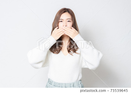 A young woman standing in front of a white wall and hiding her mouth with her hands A young woman standing in front of a white wall and hiding her mouth with her hands 73539645