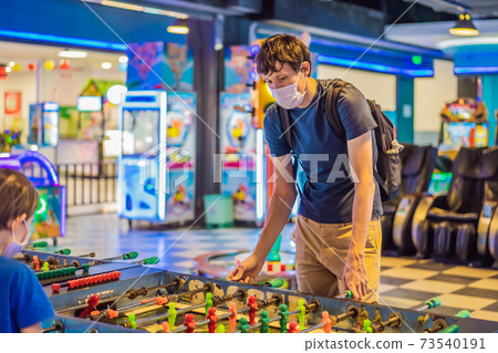 A young man wearing a medical mask during COVID-19 coronavirus playing table football indoors against a friend A young man wearing a medical mask during COVID-19 coronavirus playing table football indoors against a friend 73540191