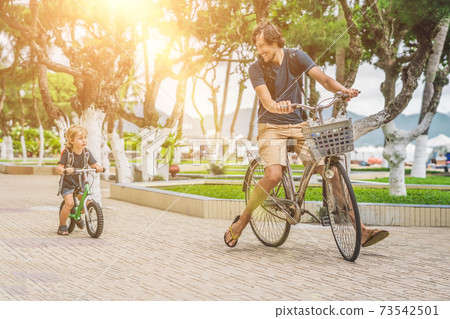 Happy family is riding bikes outdoors and smiling. Father on a bike and son on a balancebike Happy family is riding bikes outdoors and smiling. Father on a bike and son on a balancebike 73542501
