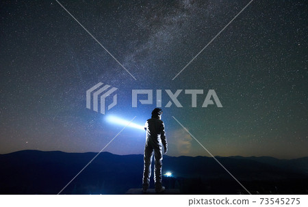 Spaceman directs a ray of light into the sky with a help of a lantern in the night in the mountains 73545275