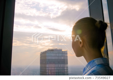 Woman looking at cityscape through window of skyscraper - sun lens flare 73549027