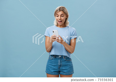 Entertained charming young european female with blond hair in casual outfit holding smartphone being satisfied with great camera and photos gasping and gazing amazed at screen over blue wall Entertained charming young european female with blond hair in casual outfit holding smartphone being satisfied with great camera and photos gasping and gazing amazed at screen over blue wall 73556301