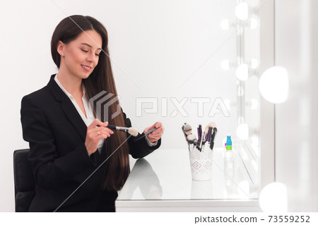 girl, make-up artist, with long dark hair in a business suit sitting at the make-up table in a bright room, office, in front of her shadows, brushes and other tools girl, make-up artist, with long dark hair in a business suit sitting at the make-up table in a bright room, office, in front of her shadows, brushes and other tools 73559252