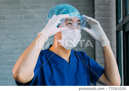 Male medical worker putting on protective gears like latex gloves, surgical face mask, bouffant cap, medical goggles at work. Asian man wearing and getting ready for working at hospital 73561026