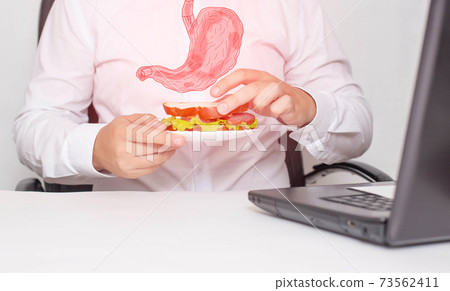A male office worker holds a sandwich in his hands during lunch. The concept of unhealthy food, snacking, stomach pain. Gastritis and heartburn A male office worker holds a sandwich in his hands during lunch. The concept of unhealthy food, snacking, stomach pain. Gastritis and heartburn 73562411